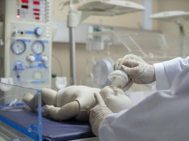 A doctor putting an oxygen mask on a newborn infant model under a radiant warmer/resuscitator unit.  clipart