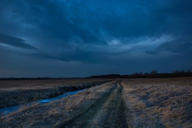 Küçük bir nehir boyunca toprak bir yol, koyu mavi bir akşam gökyüzü.