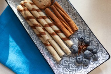 Kremalı çikolatalı gofret çubukları tarçınlı ve yaban mersinli seramik bir tabakta duruyor.