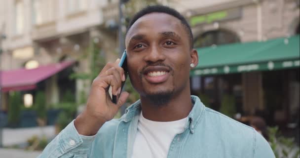 Agradable contento alegre joven barbudo de piel negra hombre en ropa de moda tiene conversación telefónica positiva cerca acogedor café de la calle en verano, cámara lenta — Vídeo de stock