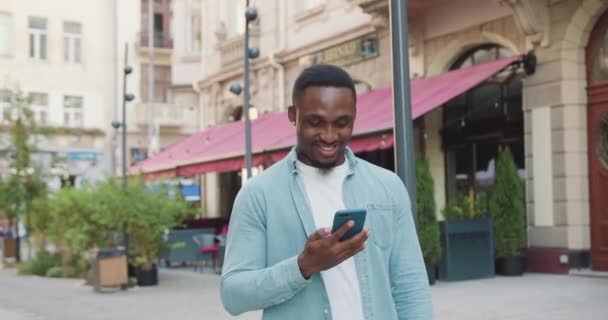 Pen smilende, ung, afroamerikansk afroamerikaner i stilige klær som går på en koselig kafe i gamlebyen og bruker mobilen sin, utsikten foran – stockvideo