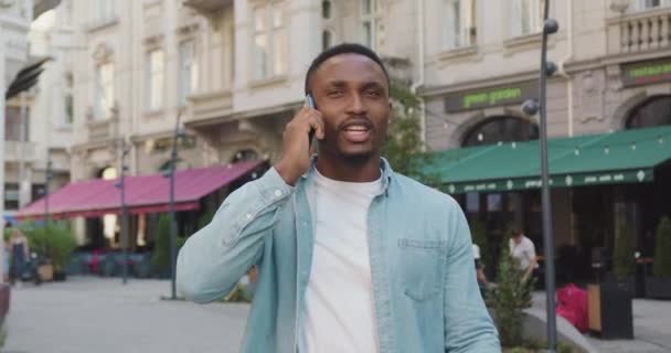 Retrato de buen aspecto alegre joven de piel negra joven en ropa casual que habla en el teléfono inteligente mientras va por la calle urbana con cafés en la parte del casco antiguo — Vídeo de stock