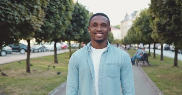 Primer plano de buen carácter despreocupado joven afroamericano alegre en ropa elegante que de pie delante de la cámara en el callejón de la ciudad con árboles verdes con una sonrisa sincera — Vídeos de Stock