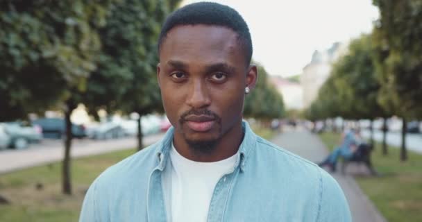 Close up of good-looking concentrated confident young black-skinned modern guy with stylish beard and in earrings which looking at camera in the middle of urban green alley — Stock Video