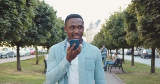 Bonito aspecto despreocupado amigable sonriente joven afroamericano en ropa casual caminando por el callejón urbano con árboles verdes y disfrutando de videollamada, vista frontal — Vídeo de stock