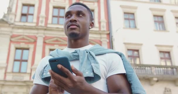 Primer plano de agradable sonriente hombre de 30 años satisfecho de piel oscura elegante con barba de moda y en pendientes que están cerca de edificios antiguos urbanos y mensajes de texto en el teléfono inteligente, 4k — Vídeos de Stock