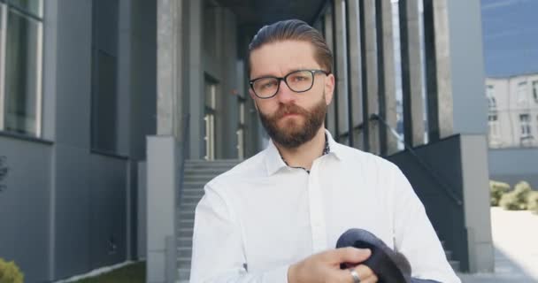 Vista frontal de atraente sério bem sucedido proposital elegante barbudo homem de negócios em óculos que posa na câmera no fundo do edifício moderno urbano — Vídeo de Stock