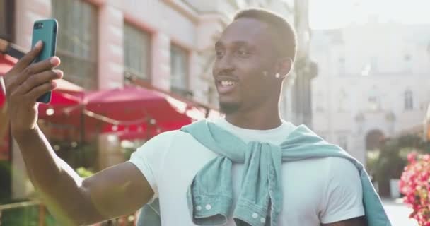 Bien parecido feliz sonriente joven afroamericano en ropa casual tiene videollamada positiva mientras está de pie cerca de acogedores cafés callejeros con hermosas flores en el día soleado — Vídeos de Stock