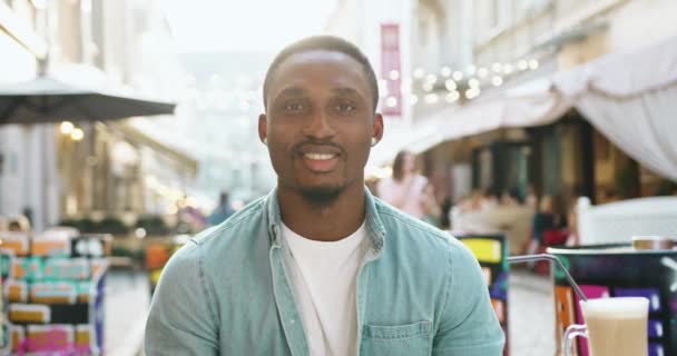 Přední pohled na dobromyslný pozitivní spokojený mladý africký Američan se stylovým vousem v neformálním oblečení, který se dívá na kameru v útulné pouliční kavárně během přestávky na kávu — Stock video