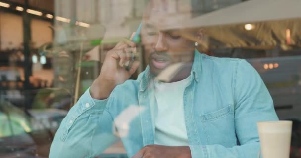 Blick durch das Fenster der Cafés, wo gut aussehende ernst selbstbewusste junge, stylische afrikanisch-amerikanische Gespräche in Zeitlupe auf dem Handy geführt werden — Stockvideo