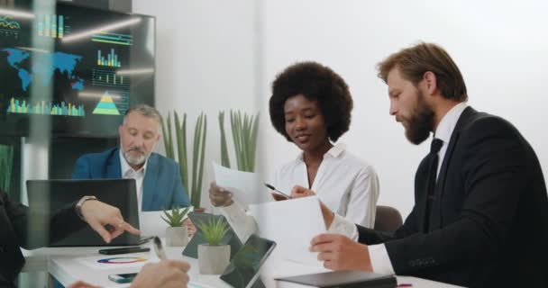 Concepto de negocio donde el hombre barbudo guapo discutiendo con su pareja de piel negra informes financieros durante la reunión en la sala de conferencias con otros miembros del grupo — Vídeos de Stock