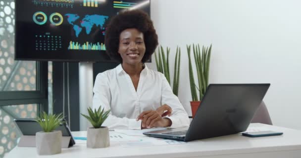 Adorable positiva alegre exitosa joven mujer de negocios de piel negra con peinado afro posa en la cámara con una sonrisa sincera en su habitación de oficina — Vídeo de stock