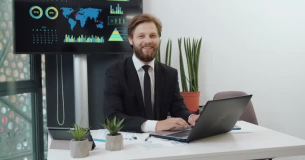 Sannolikt positiv framgångsrik självsäker skäggig VD i svart kostym sitter på sin arbetsplats i kontorsrum och tittar in i kameran med uppriktigt leende — Stockvideo