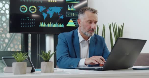 자신감 넘치는 50 세의 수염을 가진 사업가가 컴퓨터 앞에 앉아 사무실에서 사업 프로젝트를 하는 모습을 보여 줬다. — 비디오