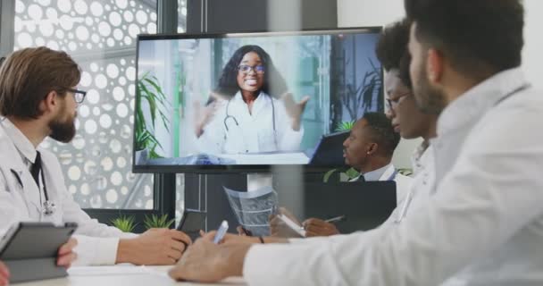 Привлекательная целеустремленная команда опытных врачей проводит онлайн видео встречу со своей коллегой на интерактивном экране — стоковое видео