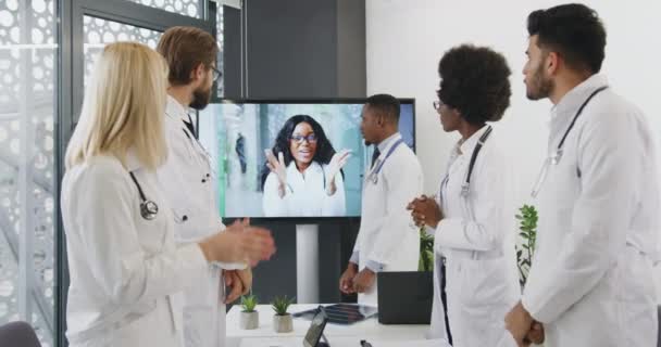 Красивая улыбающаяся темнокожая доктор приветствует свою разнообразную медицинскую команду с правом получить решение во время онлайн-конференции и поощрительные аплодисменты друг другу — стоковое видео