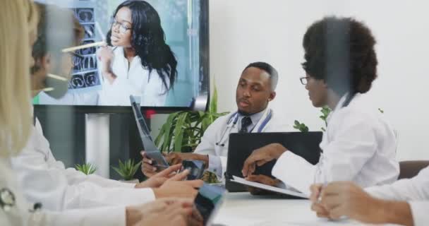 Attraente fiducioso esperto successo squadra medica diversificata che tiene una riunione in sala riunioni e consulenza per quanto riguarda i risultati della radiografia del paziente con collega femminile tramite videochiamata — Video Stock