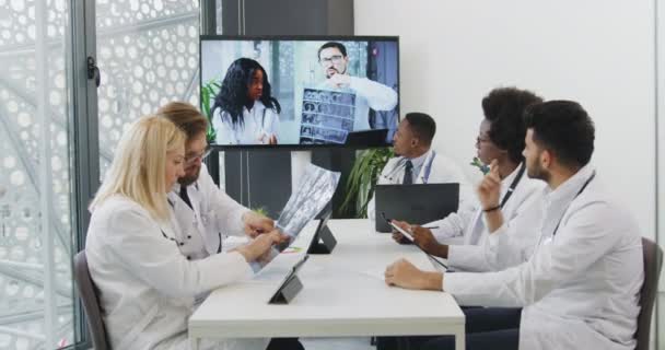 Красивые уверенные высококвалифицированные высококвалифицированные медицинские работники, сидящие за столом в офисе и имеющие онлайн видеоконференцию с международными партнерами — стоковое видео