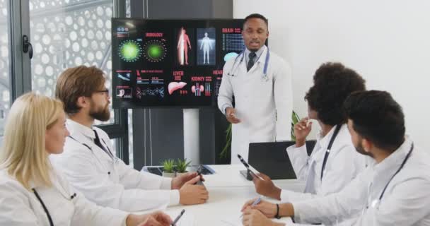 Κοντινό πλάνο του ελκυστικού επιτυχημένου πεπειραμένου μαυροδέρμου γιατρού που παρουσιάζει την ανθρώπινη εσωτερική δομή μπροστά στους διεθνείς συνεργάτες του — Αρχείο Βίντεο
