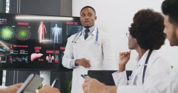 Retrato de un guapo trabajador médico de piel negra altamente calificado y confiado que sostiene una presentación sobre la estructura interna humana frente a colegas multirraciales atentos — Vídeo de stock