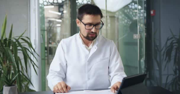 Belle confiance médecin barbu expérimenté en robe blanche regardant dans la caméra et la tenue d'une conférence en ligne avec ses collègues, au ralenti — Video