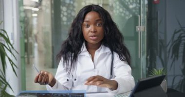 Beyaz önlüklü, kendinden emin, video konferansı düzenleyen ve hastalara danışmanlık yapan, kameraya bakan, beyaz elbiseli, siyah tenli, güzel bir kadın doktorun ön görüntüsü.