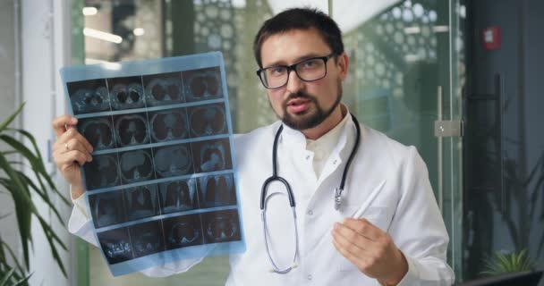 Вид спереду на доброзичливого висококваліфікованого бородатого лікаря, який проводить зустріч за допомогою відеозв'язку з партнерами та обговорює результати рентгенівського сканування — стокове відео