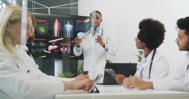 Close up of good-looking confident experienced black doctor which presenting report on digital screen on theme of internal human organs for his international colleagues in boardroom — Stock Video