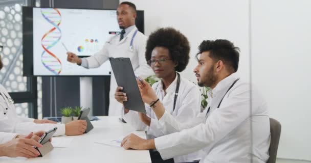 Concepto médico en el que dos médicos multirraciales confiados hombres y mujeres discuten los resultados del examen de los pacientes durante la conferencia conjunta sobre el tema del ADN con otros colegas, vista frontal — Vídeo de stock