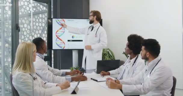Ritratto di un bel saggio medico barbuto barbuto sicuro di sé che spiega la struttura del DNA per i suoi colleghi multietnici concentrati durante l'incontro — Video Stock