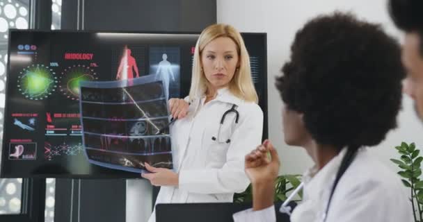 Elég magabiztos tapasztalattal rendelkező nő-orvos megbeszéli a röntgenkép eredményét a szakmai bölcs kevert faji kollégáival a tárgyalóban. — Stock videók