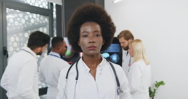 Schattige, zelfverzekerde verpleegster met een zwarte huid en Afrikaans kapsel in medische kleding met stethoscoop voor de camera, terwijl een ander lid van haar team discussieert over — Stockvideo