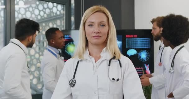 Portrait of charming confident cvalified professional female medical worker with light hair which poses on camera with crossed arms near her colleagues which discussing report on digital monitor — Stock Video