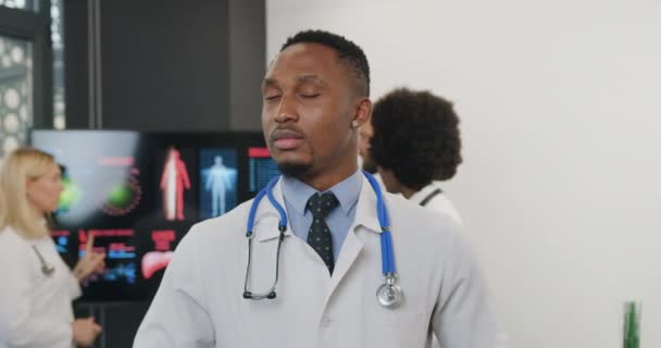 Close up of attractive confident experienced professional black-skinned male doctor in medical uniform with stethoscope which standing in front of camera on the background of his colleagues — Stock Video