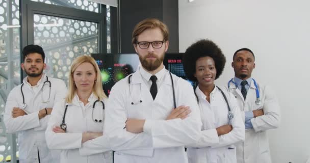 Widok z przodu dobrze wyglądającego zadowolonego, utytułowanego zespołu mieszanych pracowników medycznych w białych fartuchach ze stetoskopami, który stoi przed kamerą podczas robienia zdjęć grupowych — Wideo stockowe