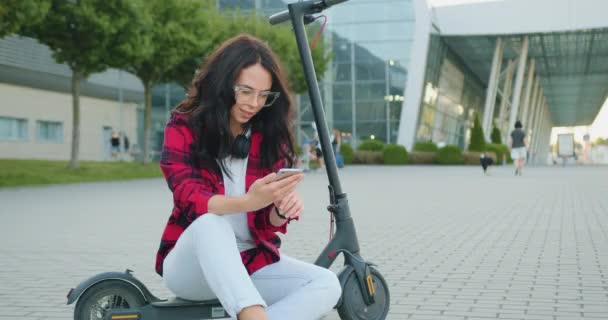 Retrato de encantador positivo satisfeito moderno elegante jovem com cabelos castanhos longos em roupas casuais que vestindo óculos e fones de ouvido em seu pescoço sentado no próprio e-scooter e usando móvel — Vídeo de Stock