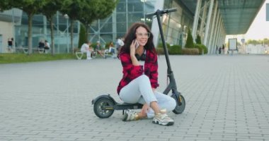 Güzel görünümlü, neşeli, kahverengi saçlı, gözlüklü ve kulaklıklı bir kadının ön görüntüsü. Bu kulaklıklar e-scooter 'da oturuyor ve büyük şehirde cep telefonuyla konuşuyor.