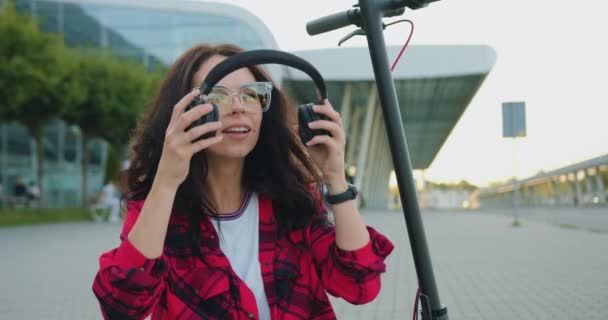 Привлекательная улыбающаяся современная довольная молодая женщина с длинными каштановыми волосами в стильной одежде в очках, надевающая наушники, чтобы слушать музыку, сидя на электронном скутере возле современного здания — стоковое видео