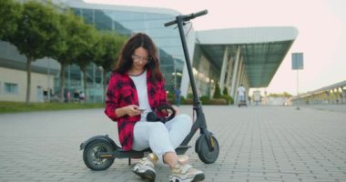 Çekici, pozitif, güler yüzlü, günlük giysiler içinde, kişisel e-scooter 'a binmiş, kulaklık takmış, şehir caddesinde, ön tarafta yürürken en sevdiği müziği dinleyen genç bir kadın.