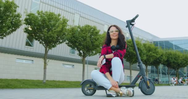 Přední pohled na atraktivní úsměv šťastný stylové mladé brunetky v módních brýlích a sluchátka na krku, která pózuje na kameře při sezení na osobní e-scooter v blízkosti městského letiště — Stock video