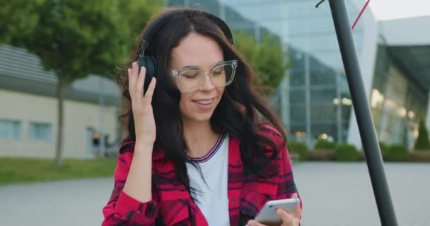 Närbild av härlig leende glad ung brunett i glasögon klädda i avslappnade kläder som lyssnar musik i hörlurar från mobil spellista på den stora urbana byggnaden bakgrund — Stockvideo