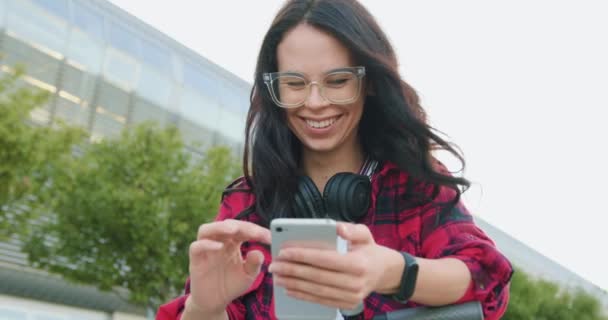 Очаровательная улыбающаяся счастливая беззаботная стильная молодая женщина с длинными каштановыми волосами в очках и в наушниках вокруг шеи, смотрящая смешные видео на телефоне во время прогулки по городской улице — стоковое видео