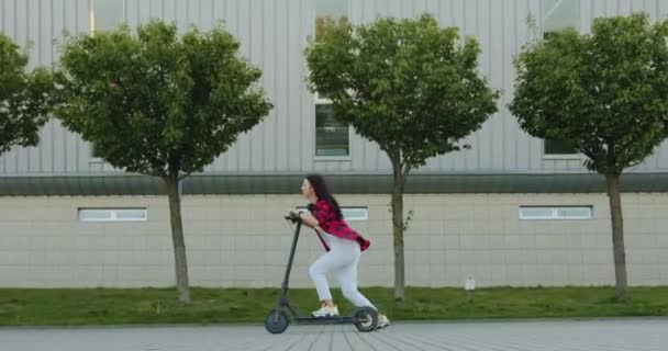 Mujer de pelo castaño alegre alegre agradable que monta en su propio e-scooter en la calle ancha con árboles verdes cerca del edificio grande contemporáneo en verano — Vídeo de stock