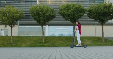 Büyük şehir binasının yanında yeşil bitkiler ve ekolojik e-scooterla gezen sıradan giysiler içindeki çekici, mutlu, genç esmer kız.