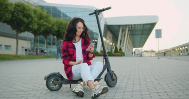 Güzel, gülümseyen, tasasız, genç esmer, gözlüklü ve modaya uygun giysileri olan şehirli modern mobiletin yanında e-scooter 'da otururken telefon çalma listesindeki şarkıların tadını çıkaran genç bir kızın ön görüntüsü.