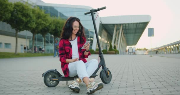 Vorderansicht der schönen lächelnden zufriedenen unbeschwerten jungen Brünetten in Brille und trendiger Kleidung, die Lieder in Kopfhörern aus der Handy-Playlist genießen, während sie auf einem E-Scooter in der Nähe von urban modern sitzen — Stockvideo