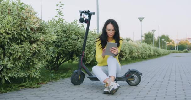 Jóképű, elegáns, figyelmes fiatal nő sárga pulóverben és világoskék farmerban ül az e-robogón, és használja a táblagépet a díszítő bokrok közelében. — Stock videók