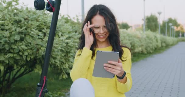 Retrato de hermosa sonriente mujer joven y elegante con gafas que utilizan su tableta mientras está sentado en su propio e-scooter cerca de hermosos arbustos verdes — Vídeo de stock