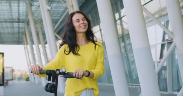 Nærbillede af charmerende smilende sorgløs glad ung brunette i gul sweater, der går med personlig e-scooter i nærheden af stor arkitektonisk bybygning om dagen – Stock-video