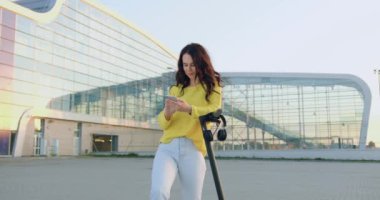 Kendine güvenen, güler yüzlü, tasasız, stil sahibi bir kız kişisel e-scooter 'ın yanında duruyor ve panoramik şehir binasının önünde cep telefonunu kullanıyor. Yavaş çekim.